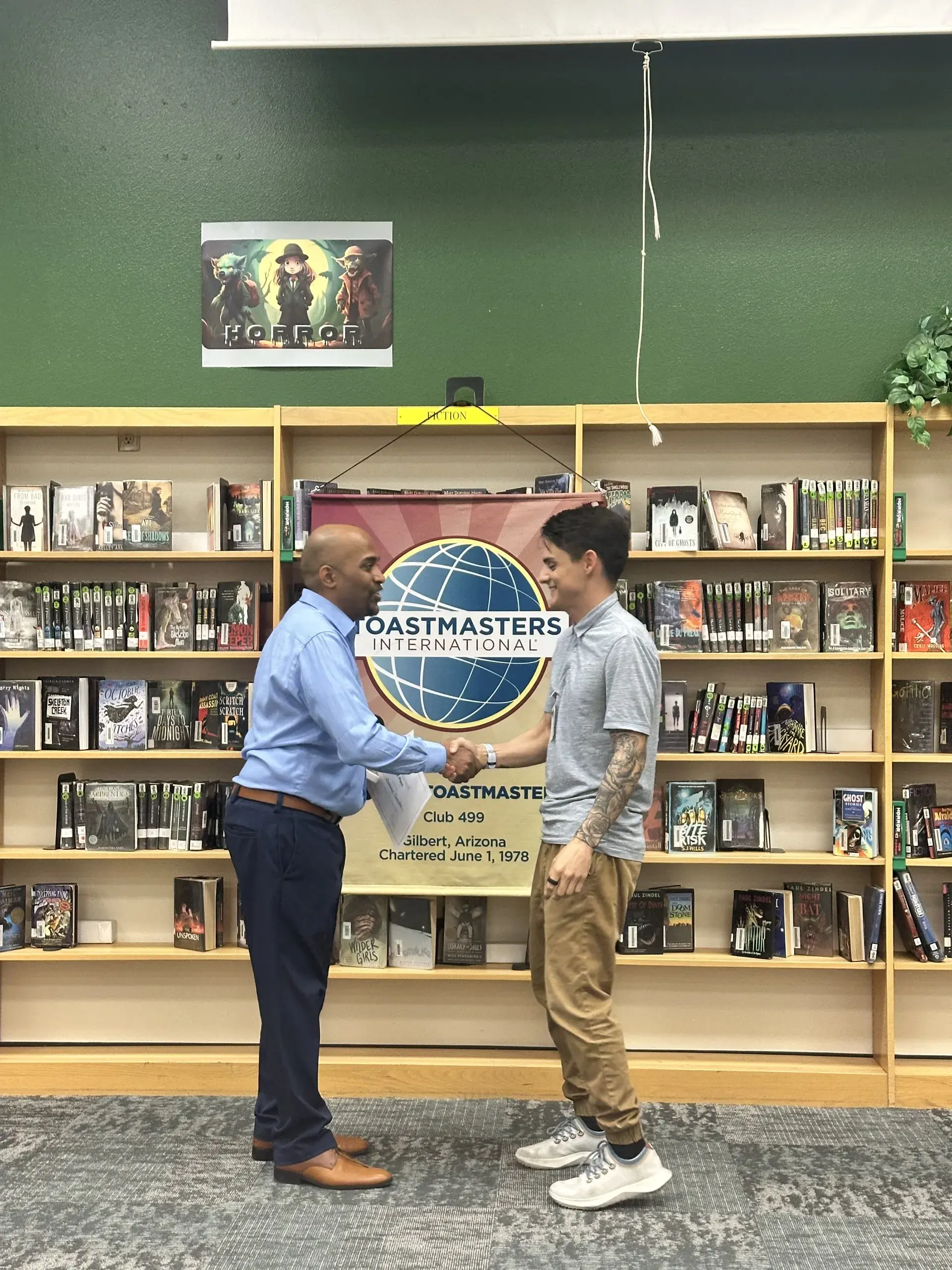 Two Gilbert Toastmasters members shaking hands in front of the club banner